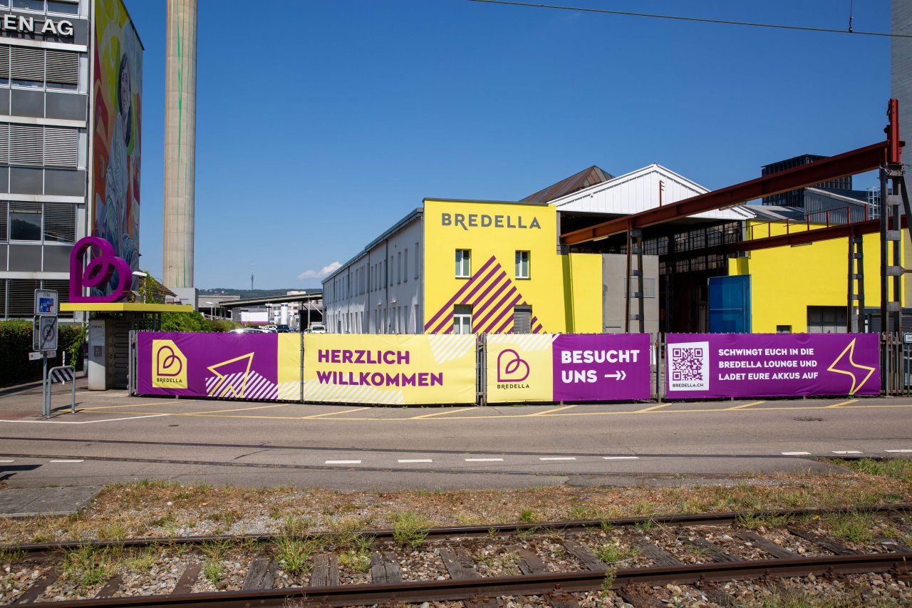 Blick vom Bahnhof Pratteln auf die alte Industriehalle, welche neu in den Bredella Farben (gelb und lila) erstrahlt. Auf dem Drehkreuzdach zum Areal steht ein grosses Bredella Herz und am Zaun hängt eine Bredella Blache.