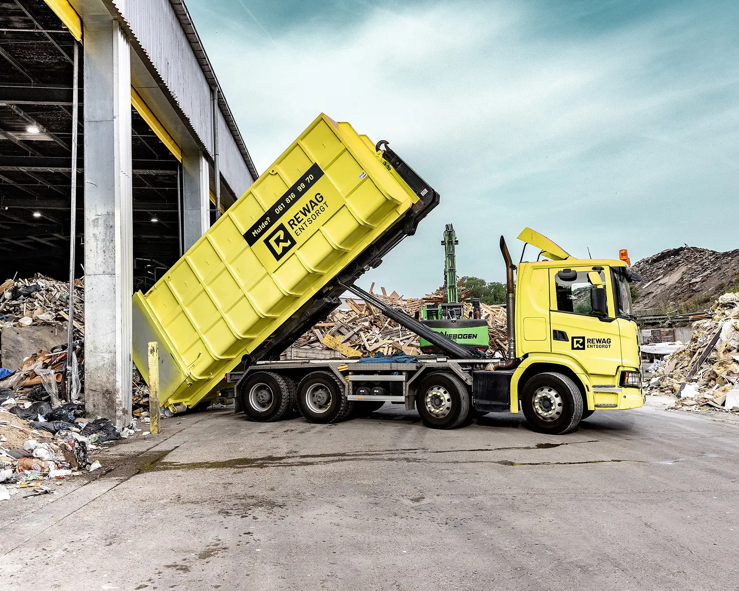 Gelber REWAG-Lastwagen kippt eine Mulde vor der offenen Sortierhalle, dahinter Bagger und Materialhaufen