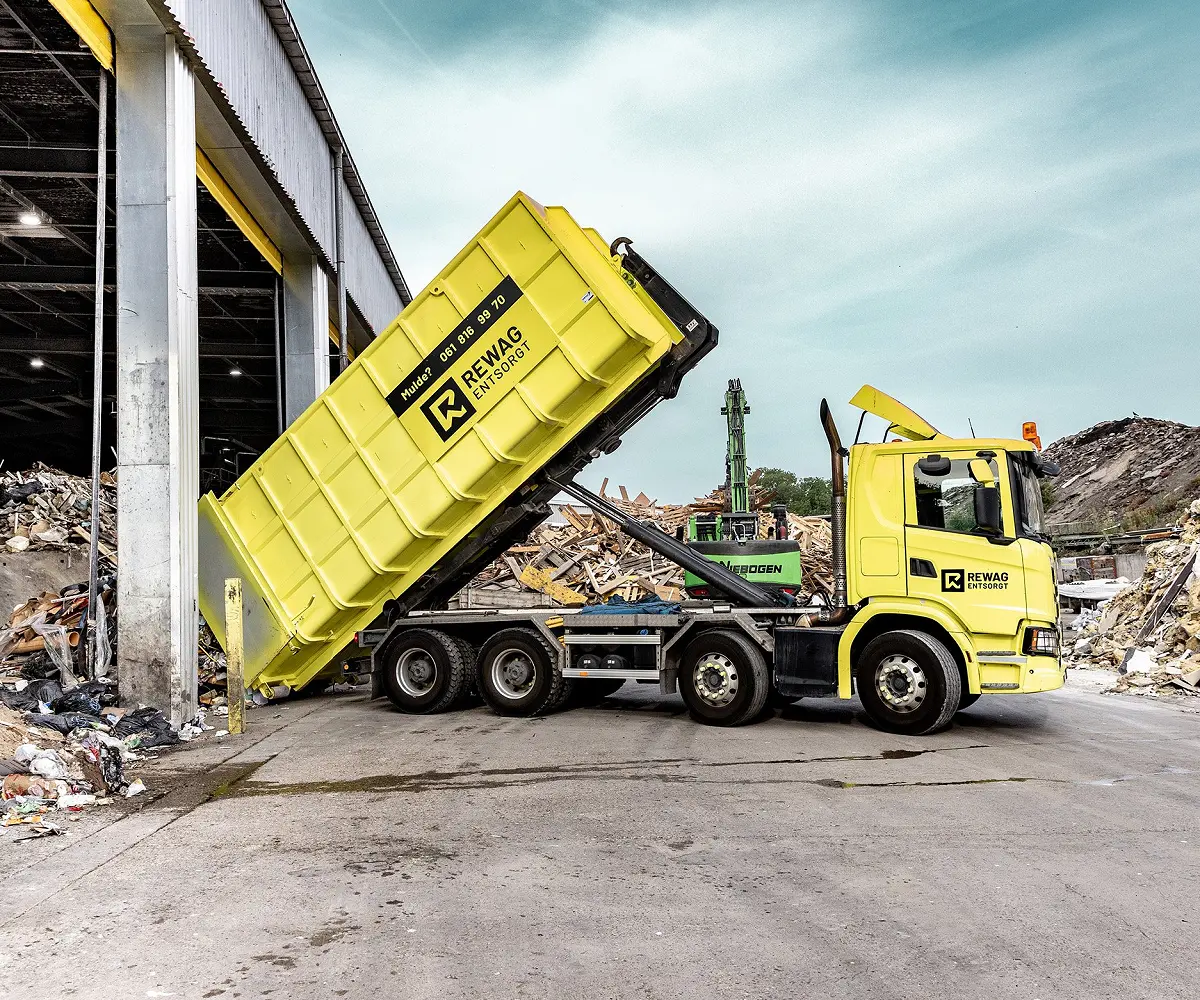 Gelber REWAG-Lastwagen kippt eine Mulde vor der offenen Sortierhalle, dahinter Bagger und Materialhaufen