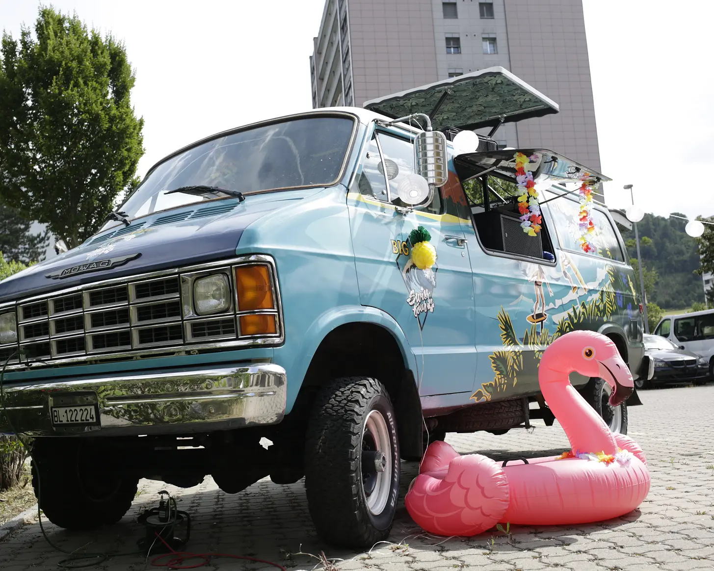 Hellblauer Van mit geöffneter Seitenklappe, davor ein rosa Flamingo-Schwimmring