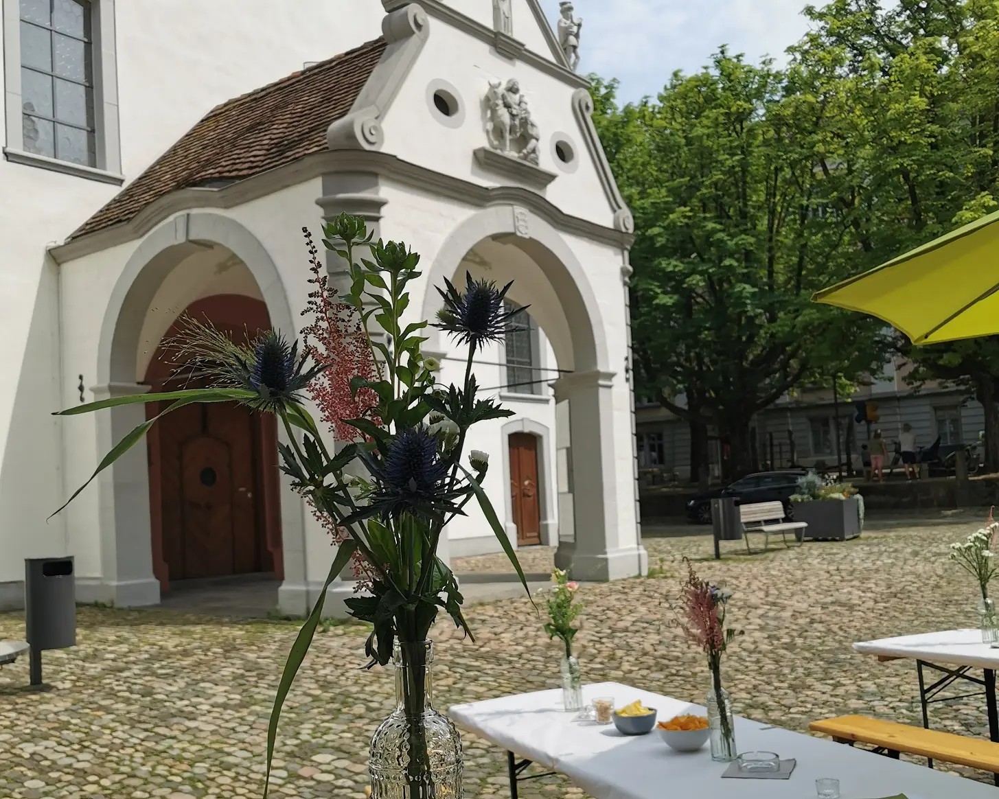 Apérotisch mit Chips und Blumenvase vor einer Kapelle, dahinter Festbänke und Sonnenschirm