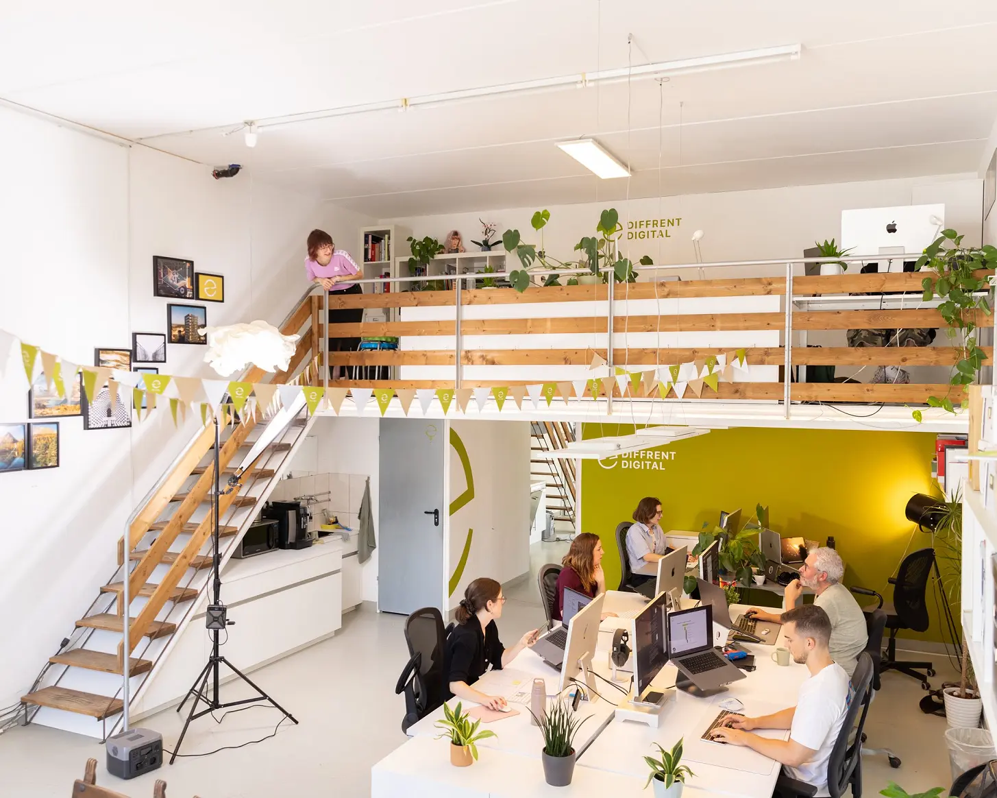 Das Atelier von der Galerie aus: Wimpelkette, Holztreppe und die Arbeitsplätze vor der gelben Wand