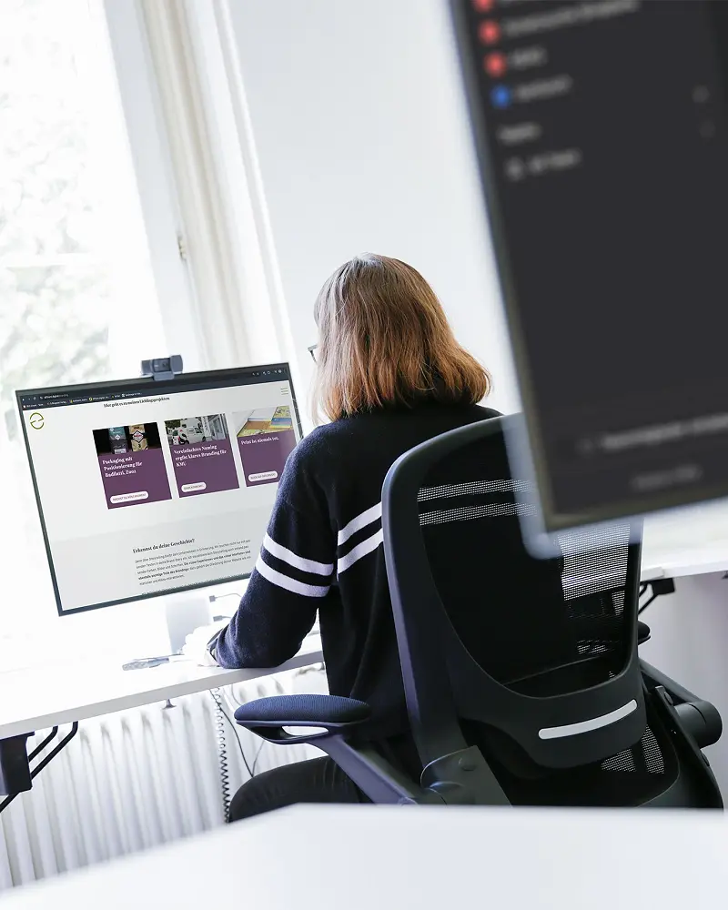 Mitarbeiterin am Schreibtisch vor zwei Bildschirmen am Fenster