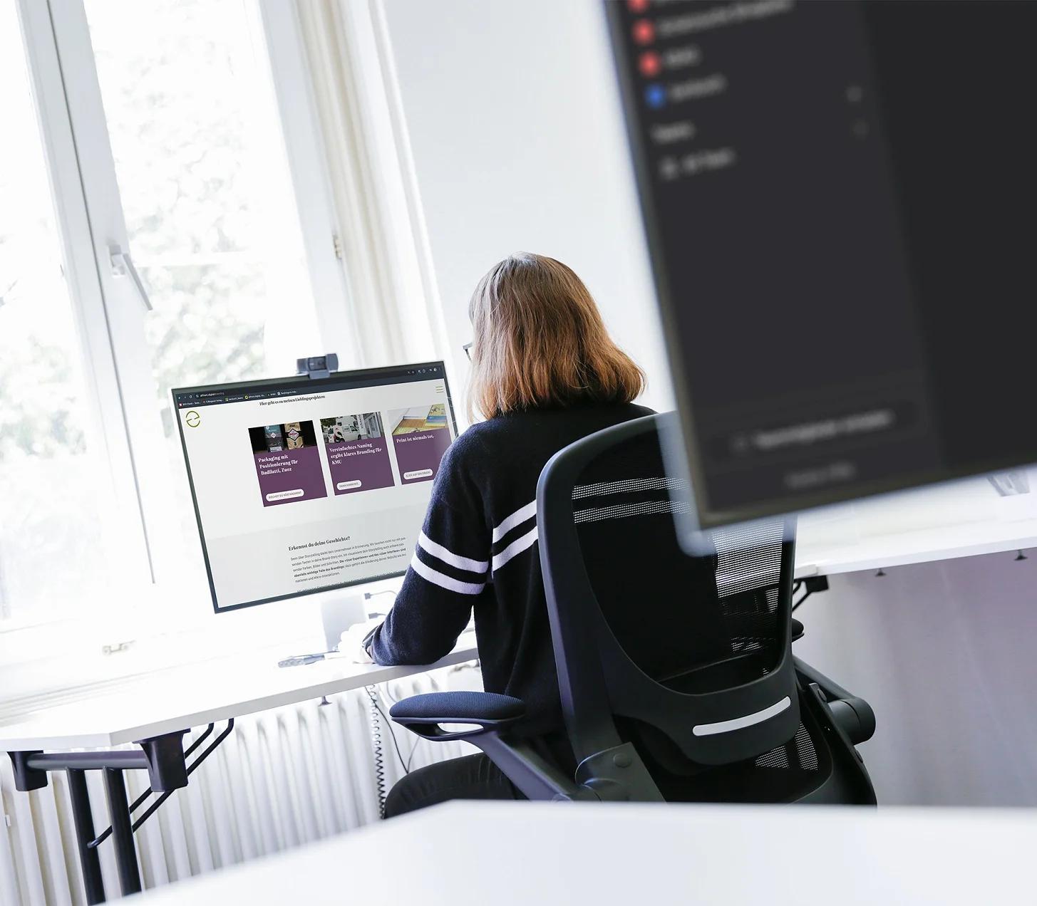 Mitarbeiterin am Schreibtisch vor zwei Bildschirmen am Fenster