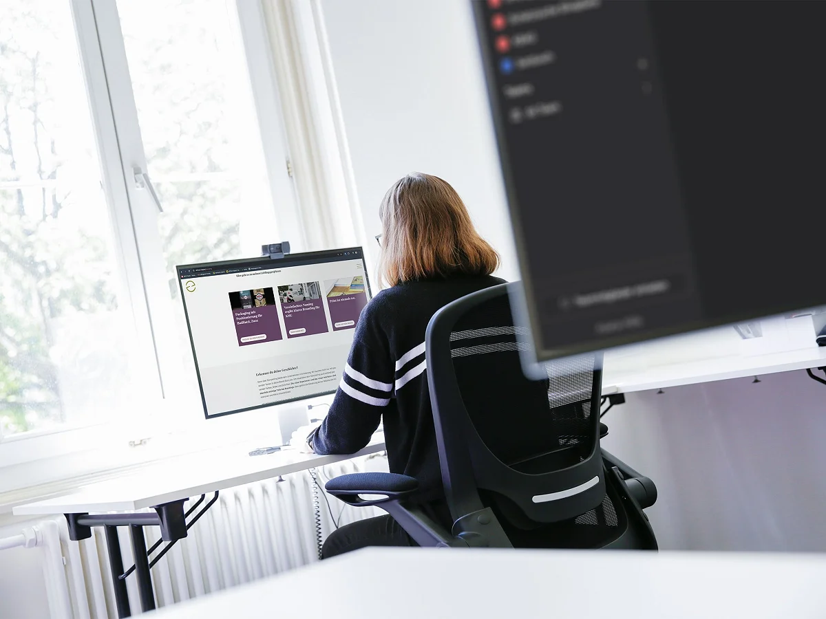 Mitarbeiterin am Schreibtisch vor zwei Bildschirmen am Fenster