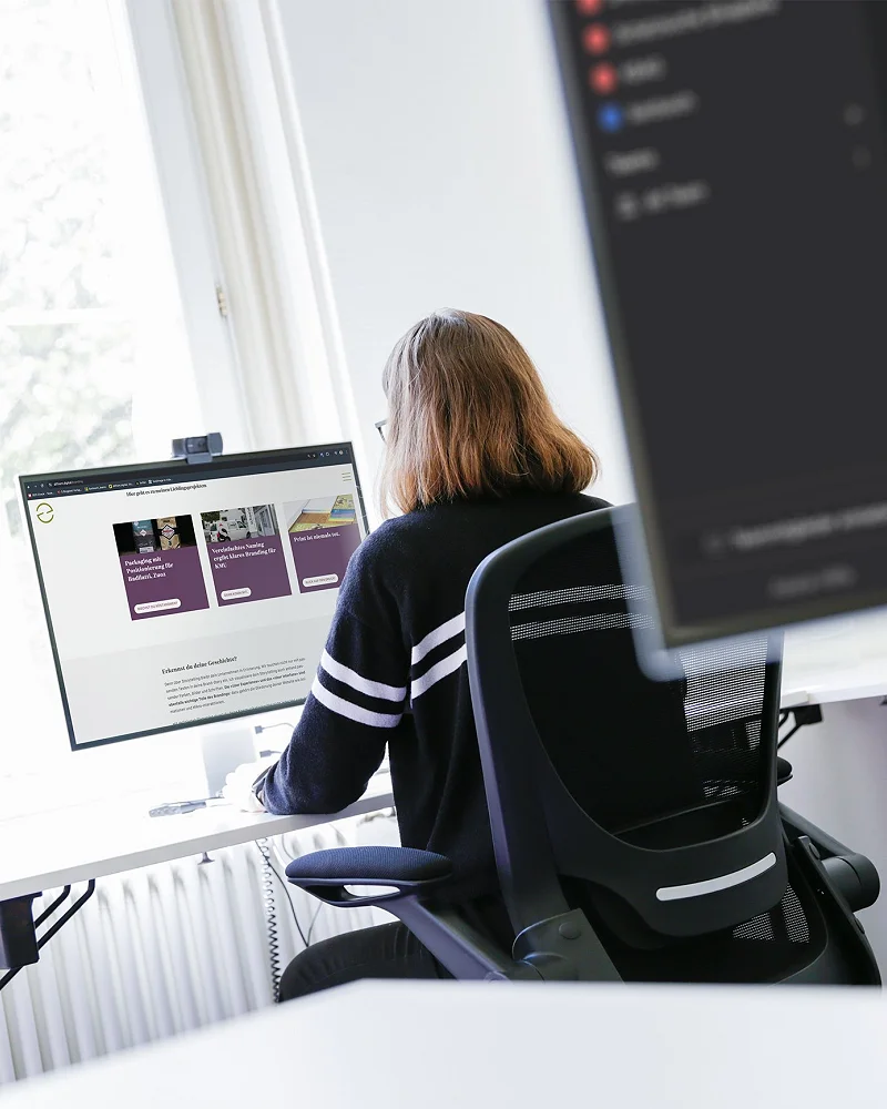 Mitarbeiterin am Schreibtisch vor zwei Bildschirmen am Fenster