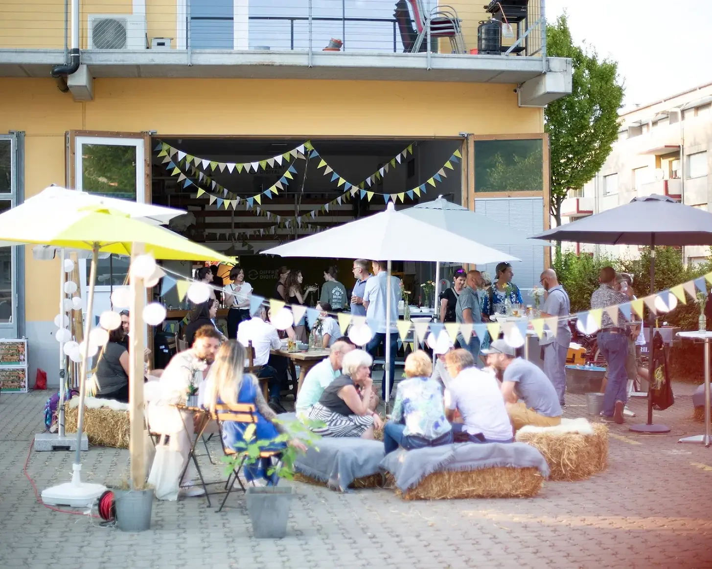 Abendstimmung am Fest: Sonnenschirme, Lichterketten und Gäste auf Strohballen vor dem Büro