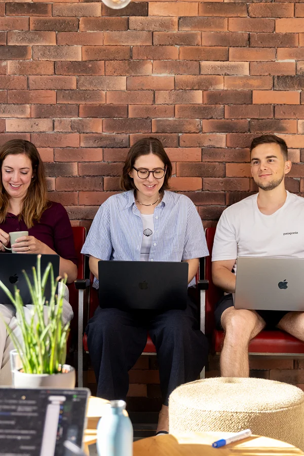Drei Mitarbeitende mit Laptops vor der Backsteinwand, rechts im Vordergrund Anita