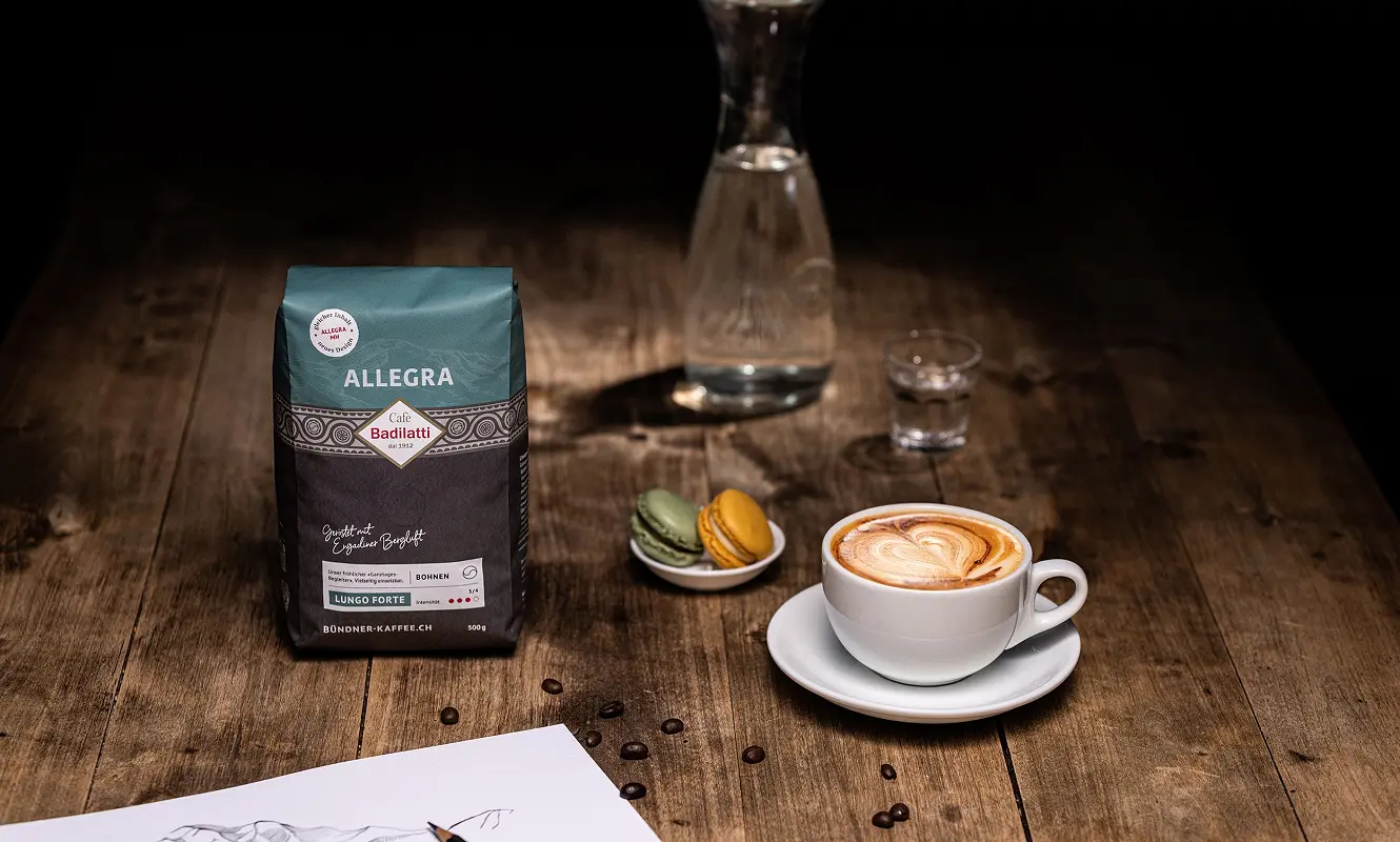 Packung «Allegra» neben Cappuccino, Macarons und einer Bergzeichnung auf dem Holztisch