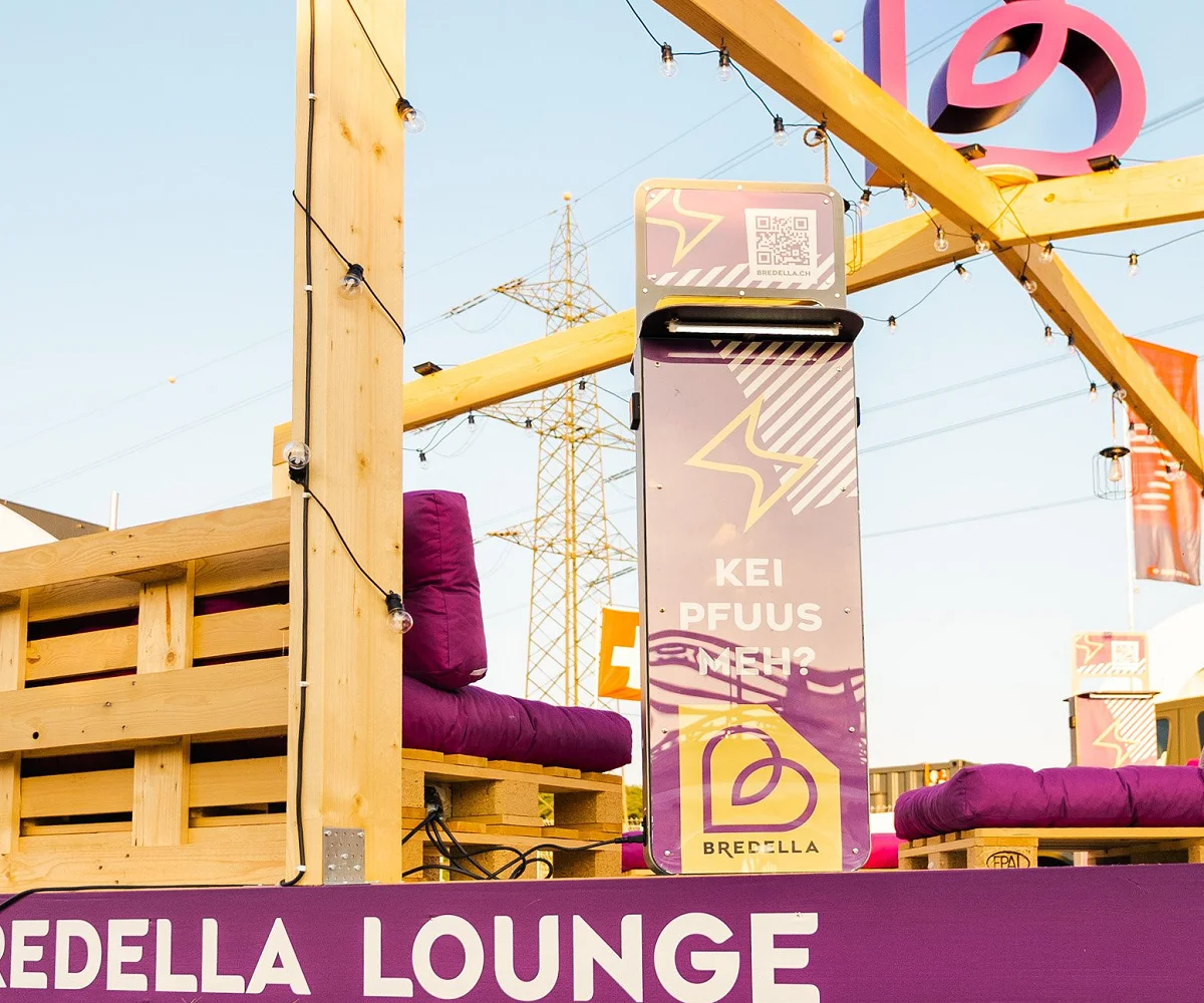 Holzrahmen der Bredella Lounge mit Signet, davor die Stele «Kei Pfuus meh?» und die violette Sockelblende