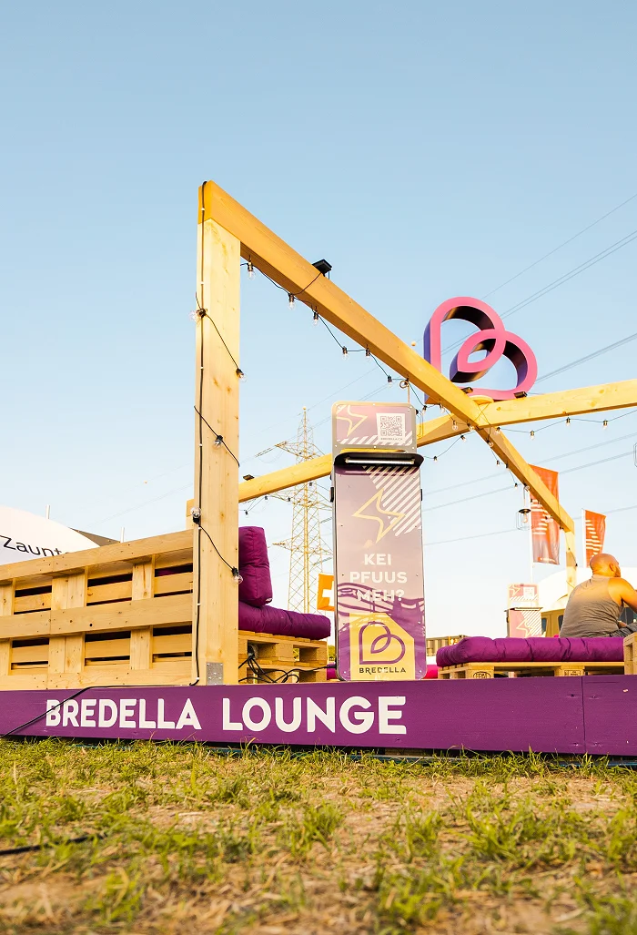 Holzrahmen der Bredella Lounge mit Signet, davor die Stele «Kei Pfuus meh?» und die violette Sockelblende