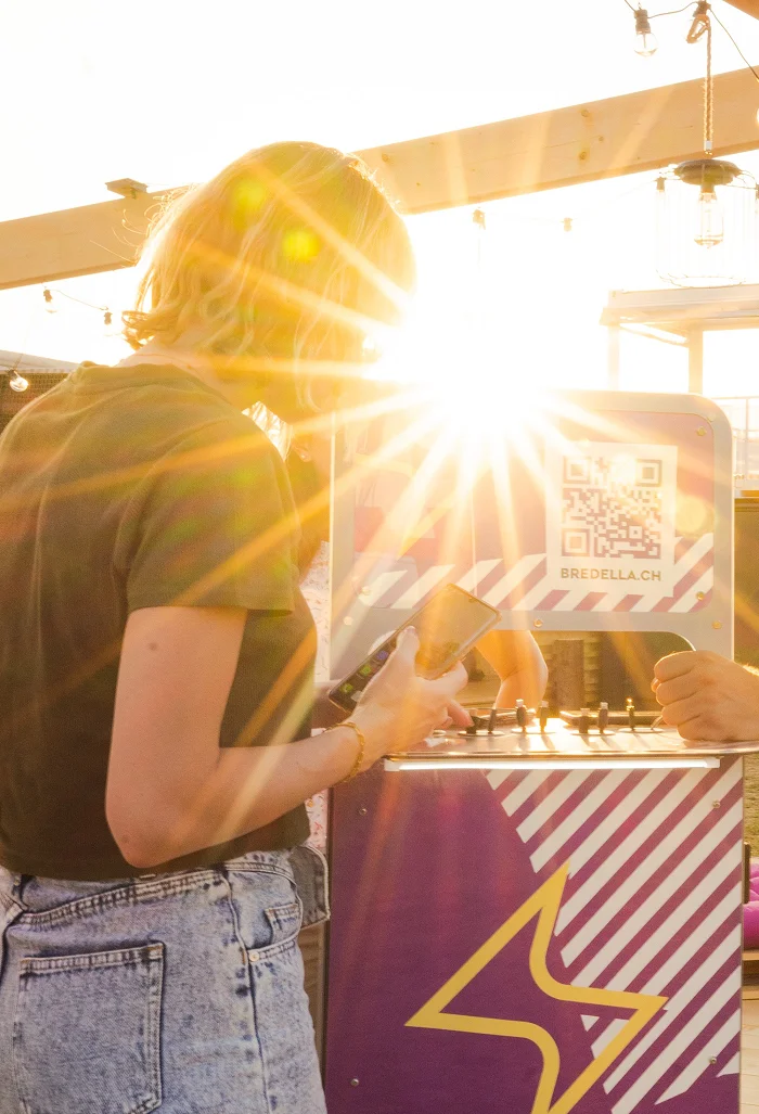 Besucherin an der Ladestation «Kei Pfuus meh?» der Bredella Lounge im Gegenlicht, auf der Stele der QR-Code zu bredella.ch