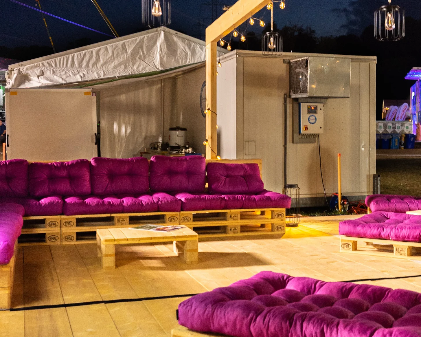 Bredella Lounge bei Nacht: Lichterketten und Hängelampen über den Palettenmöbeln, das Signet leuchtet violett