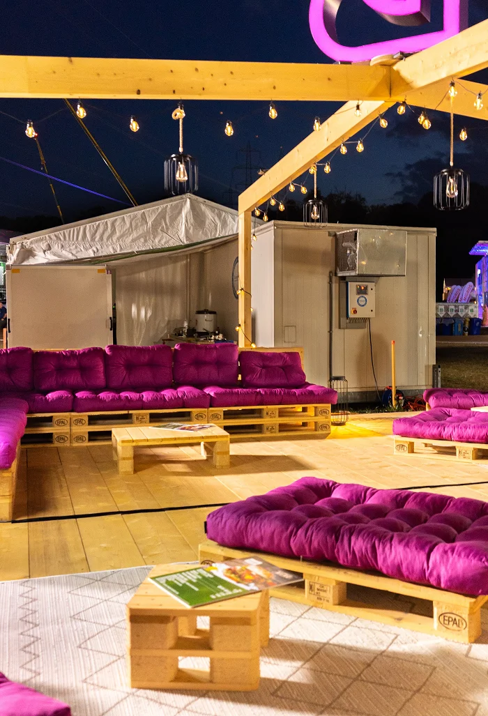 Bredella Lounge bei Nacht: Lichterketten und Hängelampen über den Palettenmöbeln, das Signet leuchtet violett