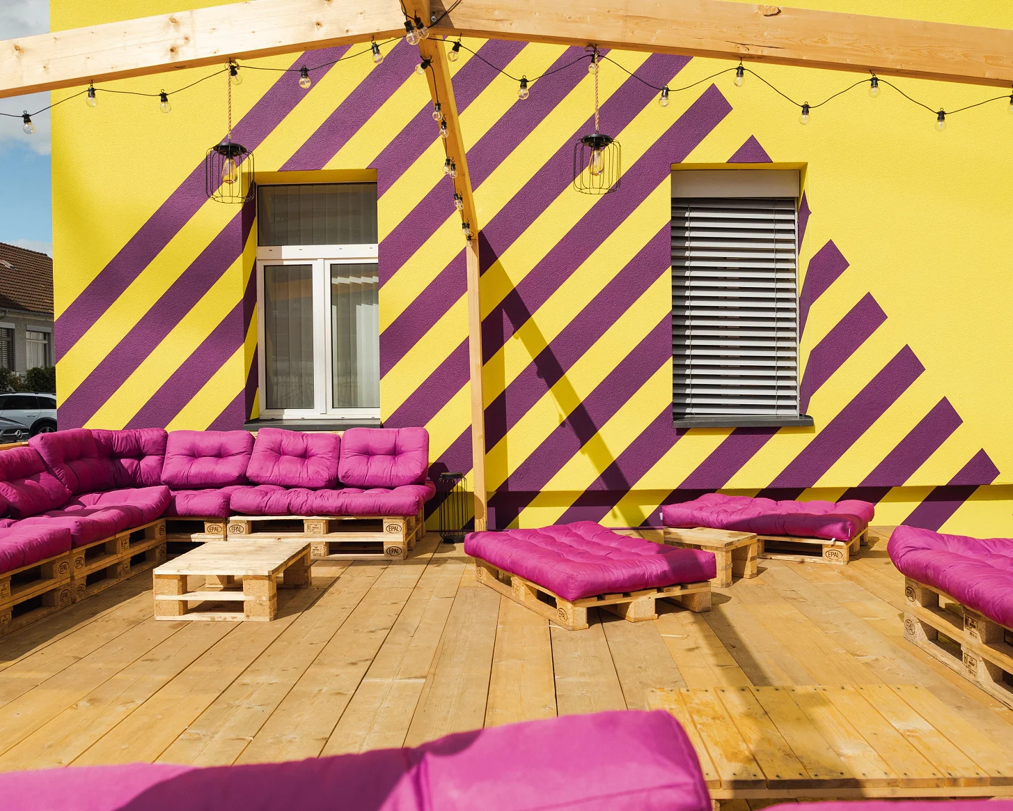 Palettenmöbel mit violetten Kissen auf einem Holzdeck vor der gelb-violett gestreiften Bredella-Fassade