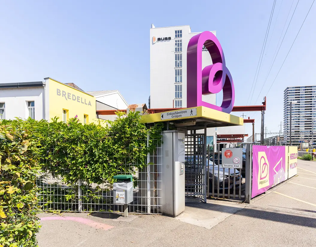 Arealeingang mit dem Bredella-Signet als Skulptur über dem Zugang, dahinter das Buss-Hochhaus