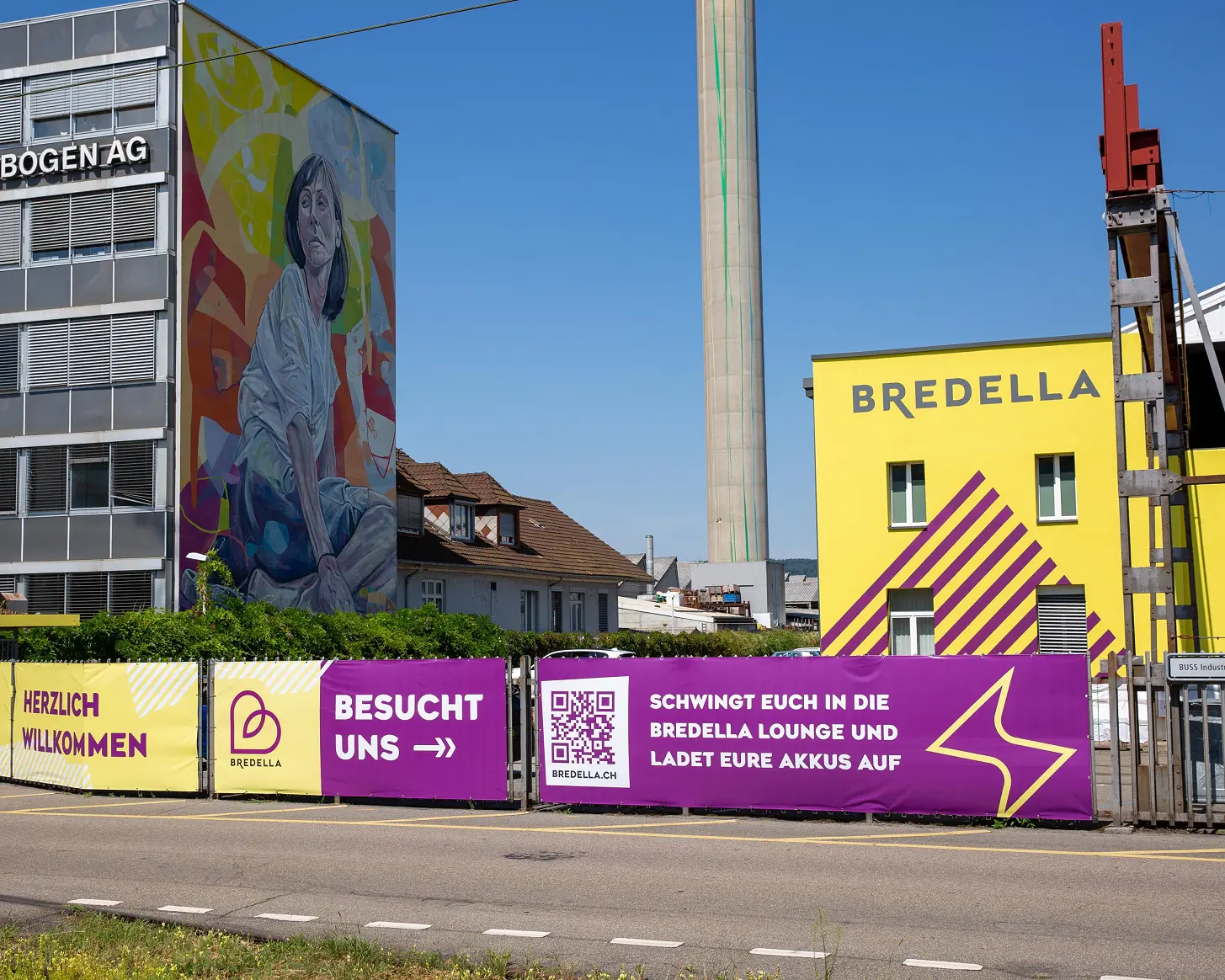 Bauzaun-Banner vor der gelben Bredella-Halle, links das Wandbild an der Rohrbogen AG