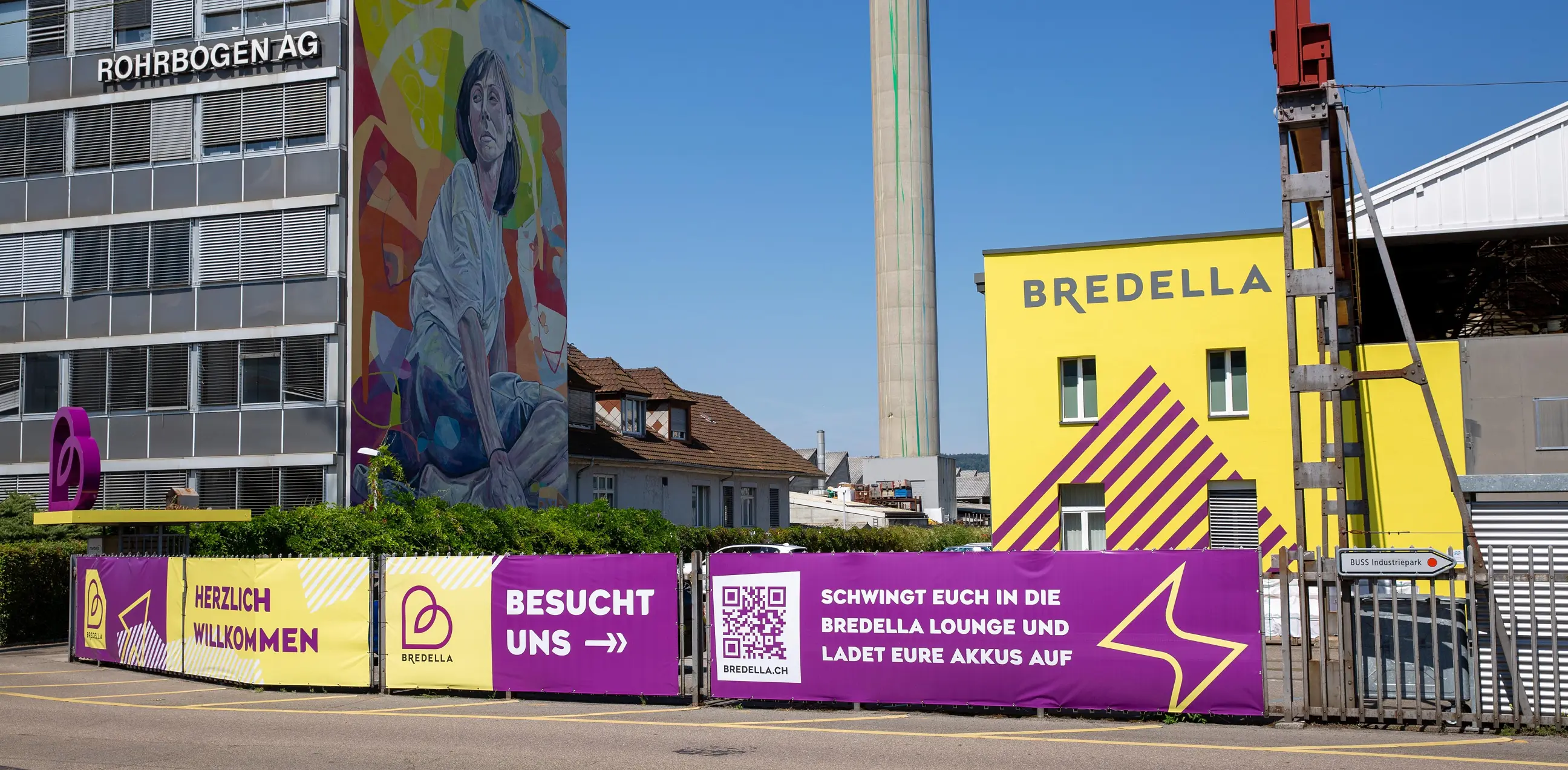Bauzaun-Banner vor der gelben Bredella-Halle, links das Wandbild an der Rohrbogen AG