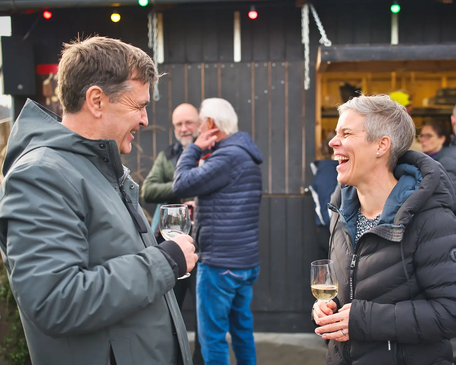 Zwei Gäste lachen mit Weingläsern vor der dunklen Holzfassade des Bredella Village. Foto Marc Gilgen