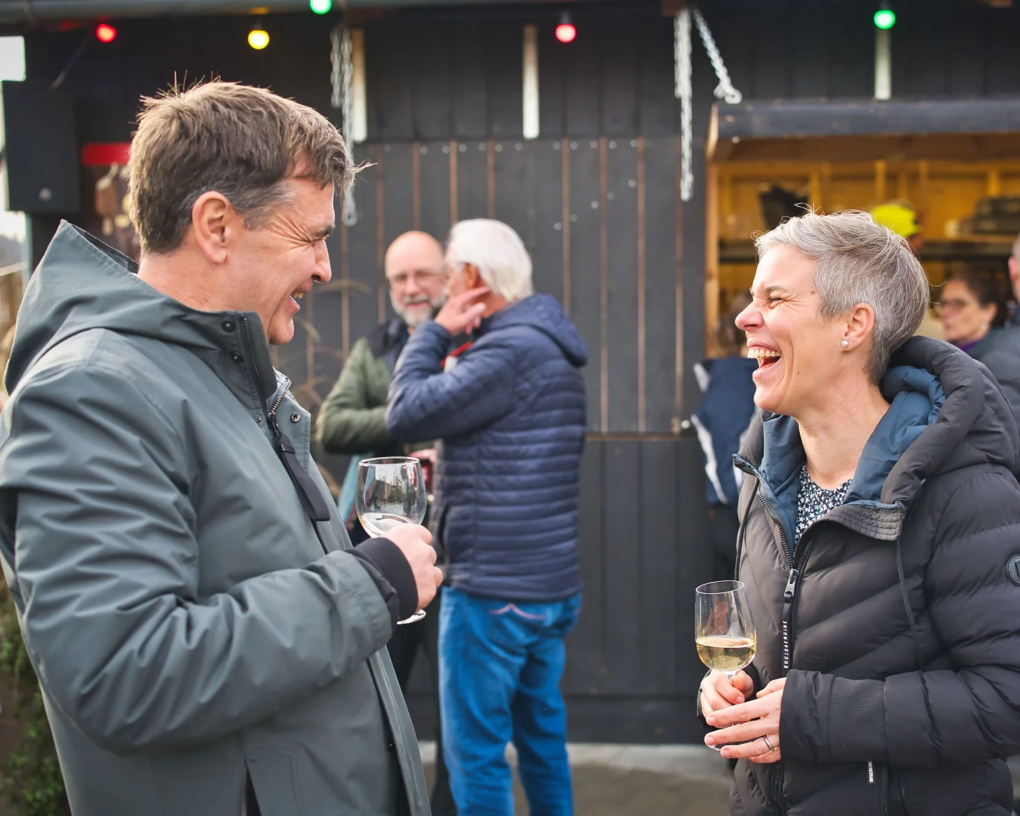 Zwei Gäste lachen mit Weingläsern vor der dunklen Holzfassade des Bredella Village. Foto Marc Gilgen