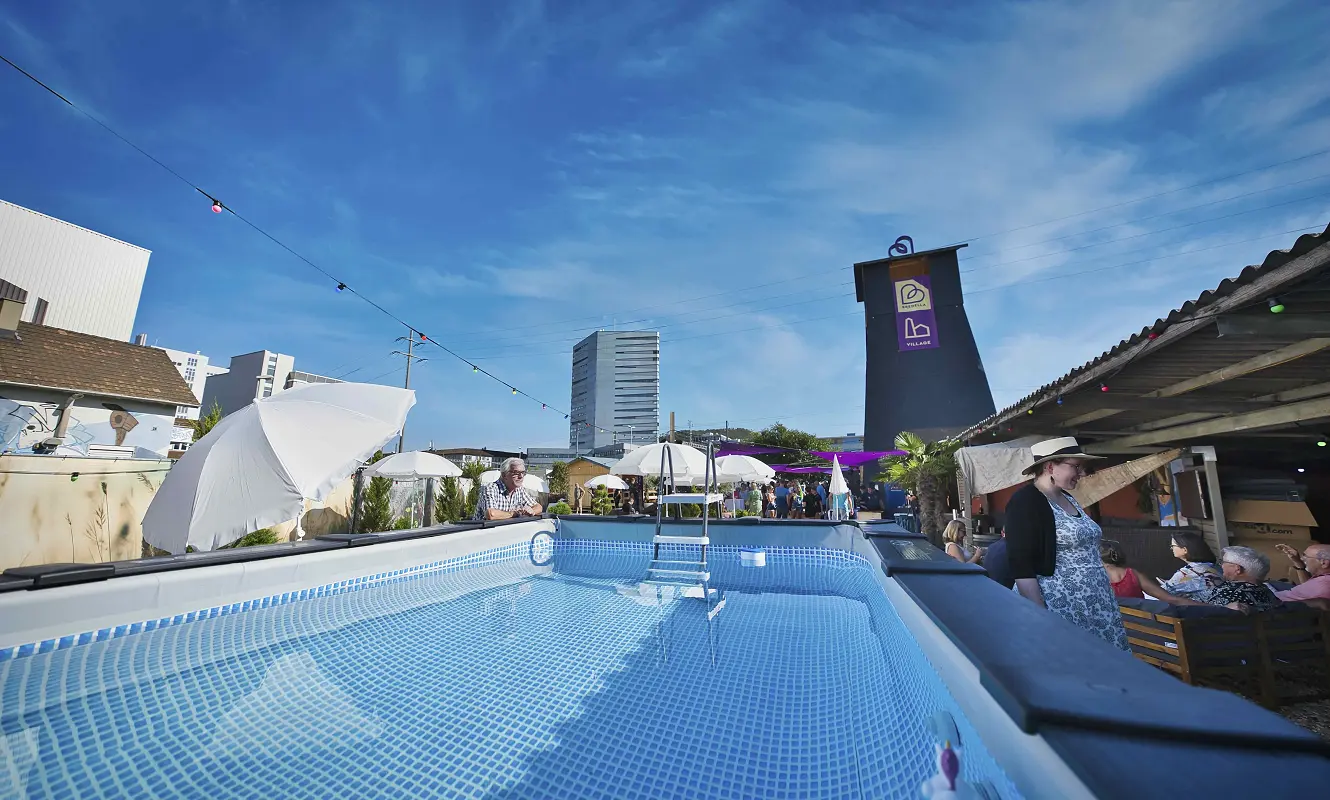 Pool im Bredella Village mit Sonnenschirmen, dahinter der Salzbohrturm mit dem Village-Banner. Foto Marc Gilgen