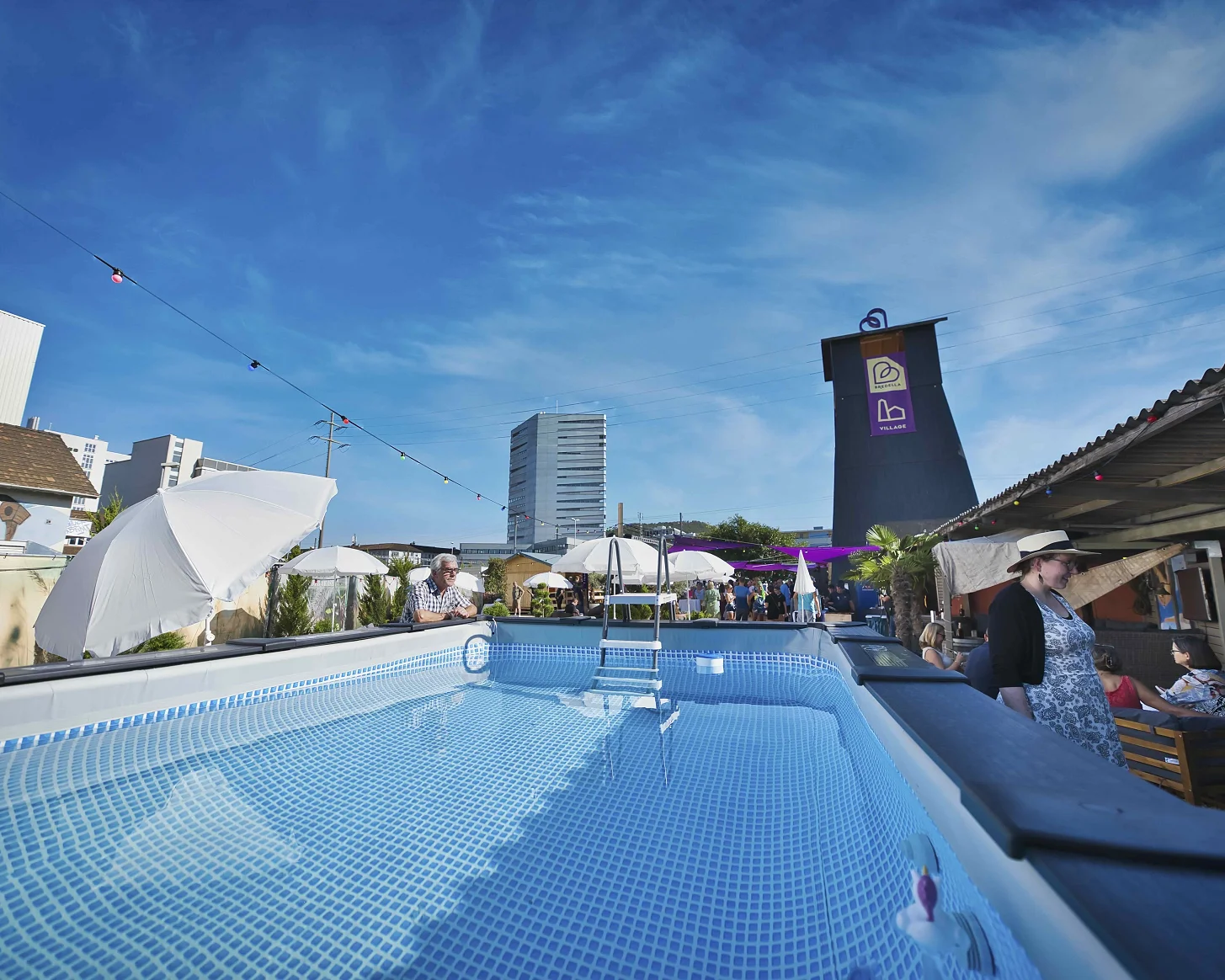 Pool im Bredella Village mit Sonnenschirmen, dahinter der Salzbohrturm mit dem Village-Banner. Foto Marc Gilgen