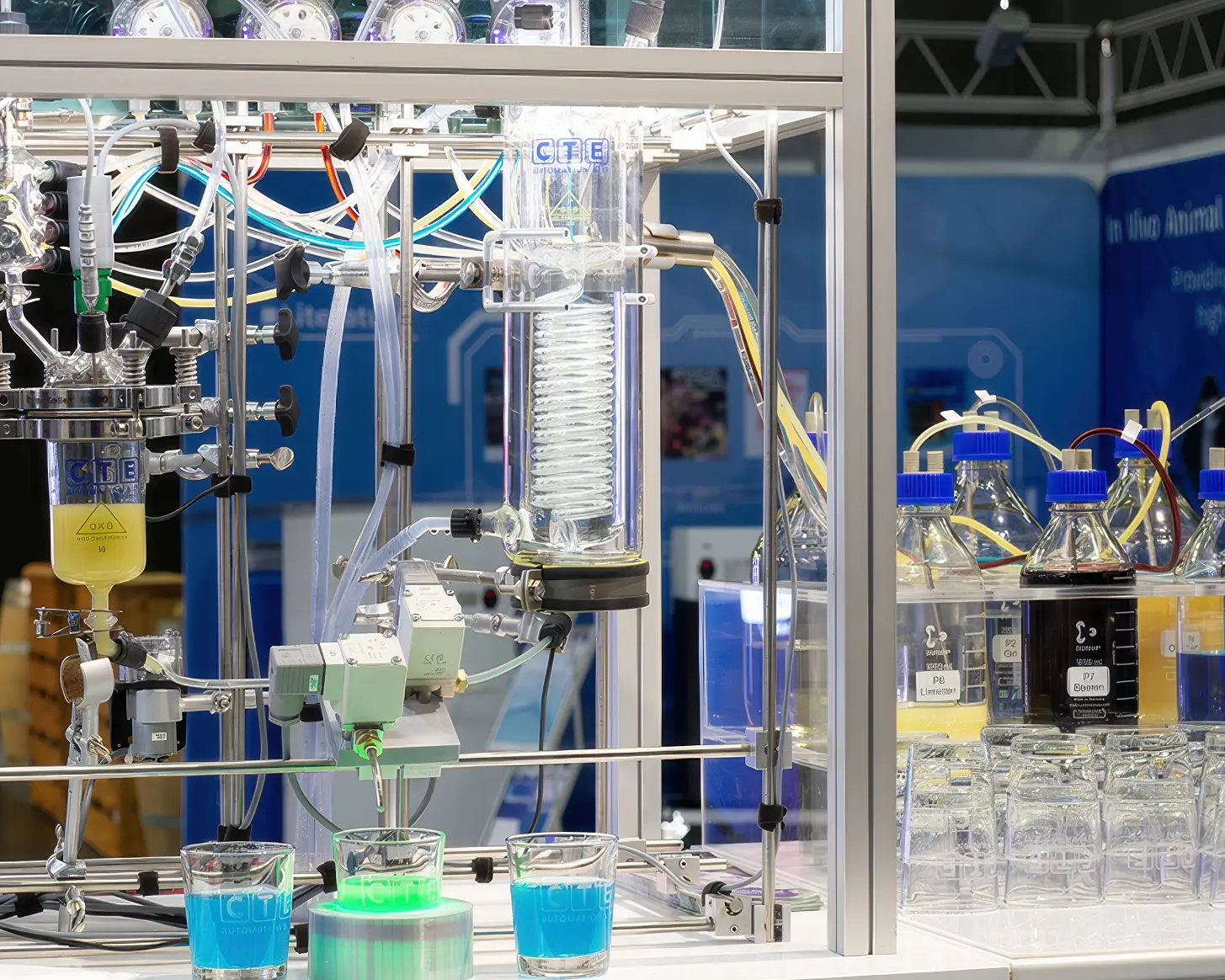 Die Cocktailmaschine am CTE-Messestand aus der Nähe: Laboranlage aus Glas und Aluminium mit Reaktor, Kühler, Ventilen und Schläuchen, davor drei Gläser mit blauem und grünem Getränk