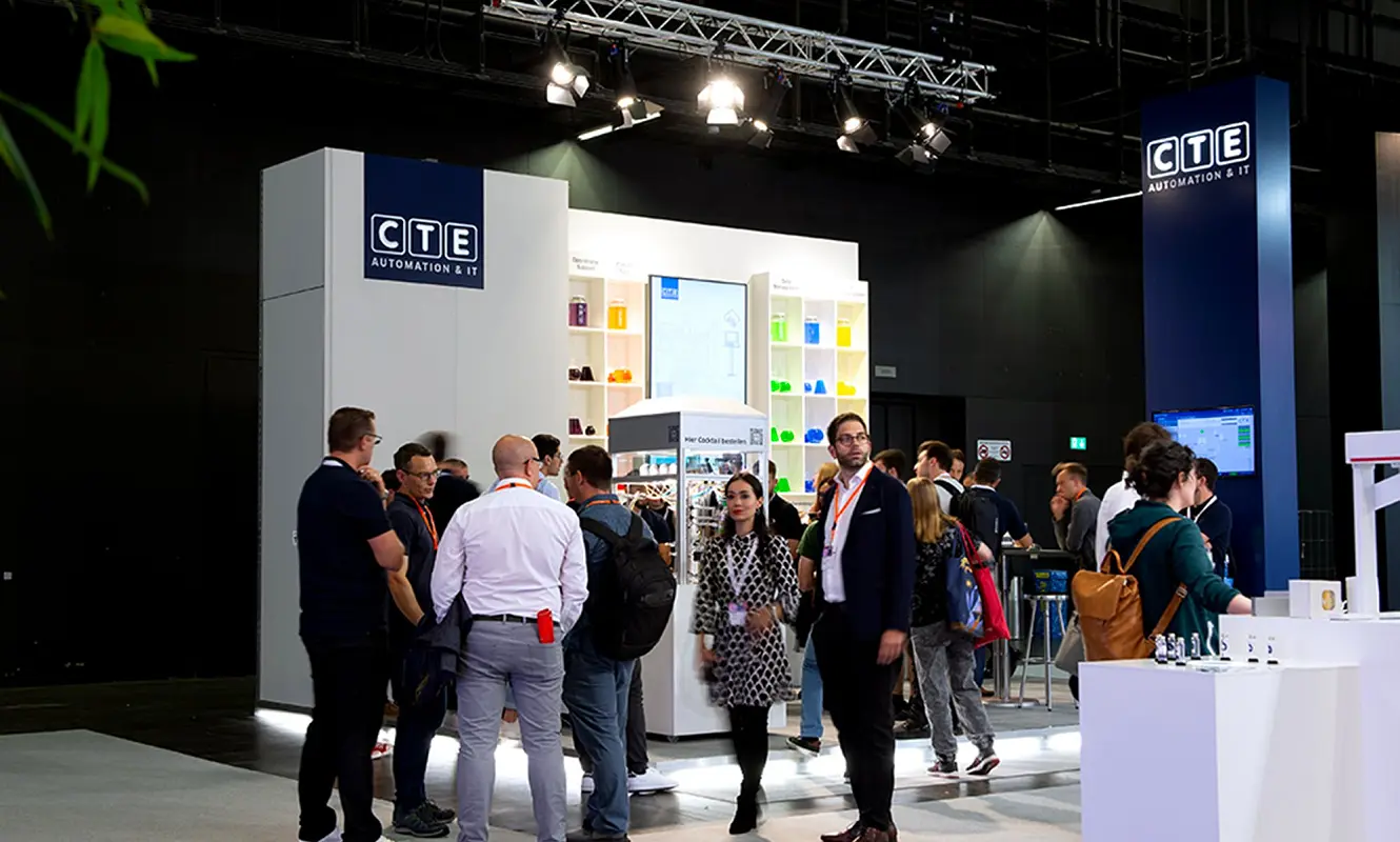 Betrieb am CTE-Stand an der Ilmac 2023: Besucherinnen und Besucher stehen vor der Regalwand und dem Cocktailkubus, rechts der blaue Turm mit dem Logo