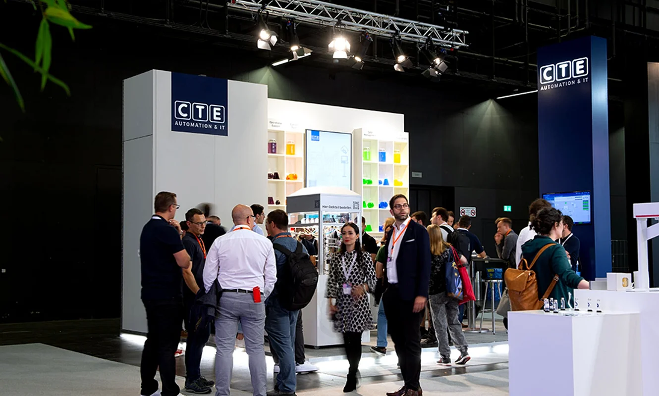 Betrieb am CTE-Stand an der Ilmac 2023: Besucherinnen und Besucher stehen vor der Regalwand und dem Cocktailkubus, rechts der blaue Turm mit dem Logo