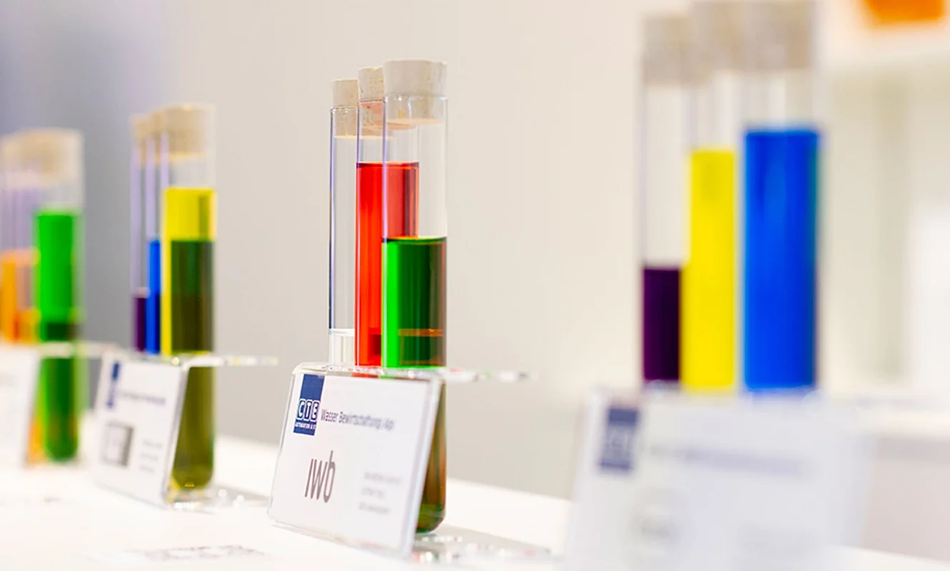 Referenzprojekte als Reagenzgläser: farbige Flüssigkeiten in Glasröhrchen auf Kartätzchen mit Kundenlogo, vorne die Wasserbewirtschaftung für iwb
