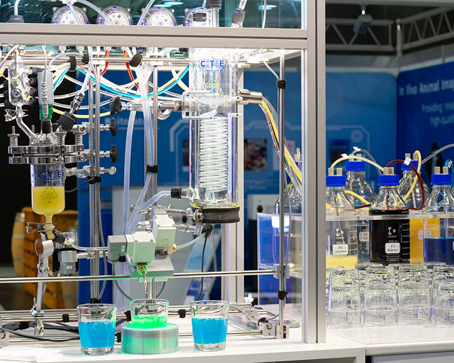 Die Cocktailmaschine am CTE-Stand aus der Nähe: Laboranlage mit Reaktor, Kühler, Ventilen und Schläuchen, davor drei Gläser mit blauem und grünem Getränk