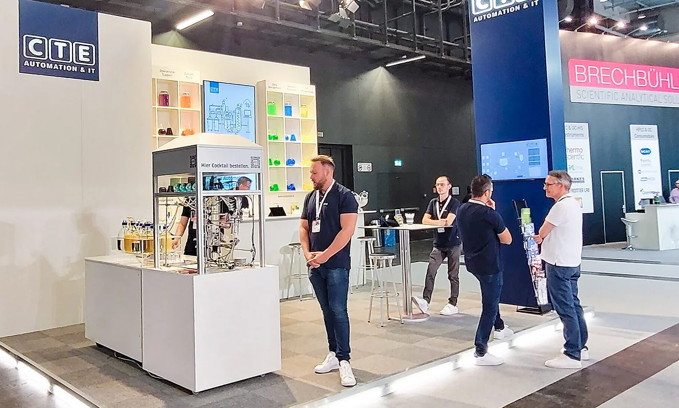 CTE-Stand von der Seite: die Cocktailmaschine auf weissem Sockel mit dem Hinweis «Hier Cocktail bestellen», dahinter Regalwand, Stehtisch und blauer Turm