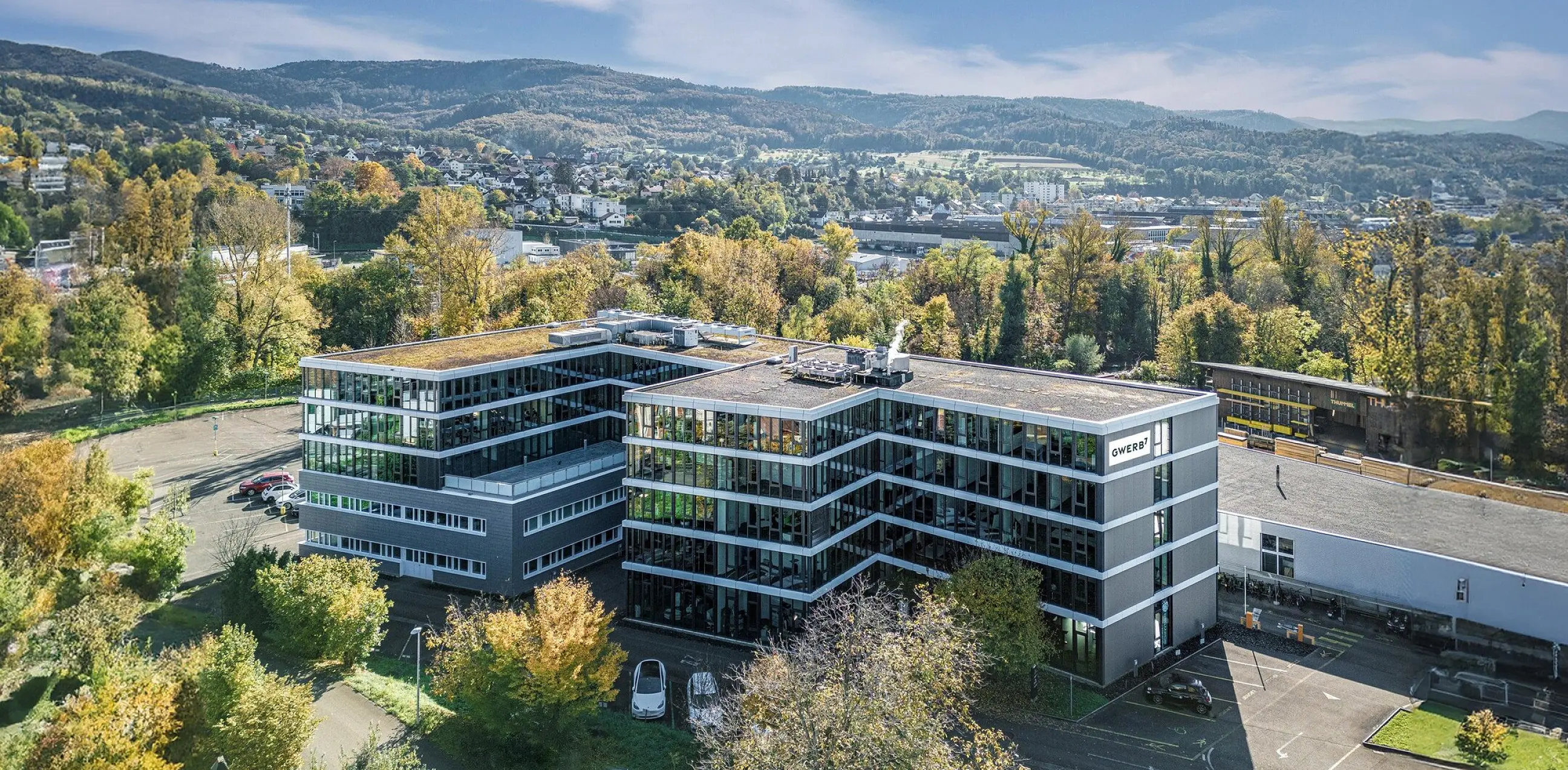 Drohnenaufnahme des GWERB7 in Aesch mit Fassadenschriftzug, begrüntem Dach und Blick über das Birstal