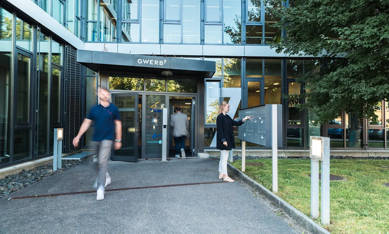 Eingang des GWERB7 mit Glasfront und Schriftzug über der Tür, davor Passanten und eine Frau am Infoschild