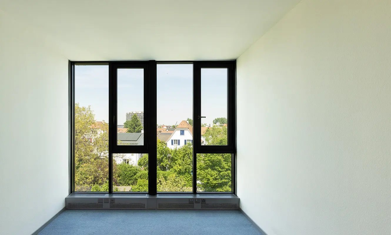 Leeres Einzelbüro im 3. OG mit bodentiefer Fensterfront, blauem Teppich und Blick über die Dächer von Aesch