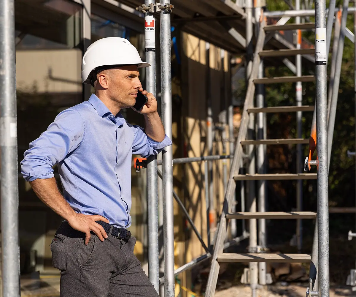 Person mit weißem Helm telefoniert auf einer Baustelle neben dem Gerüst