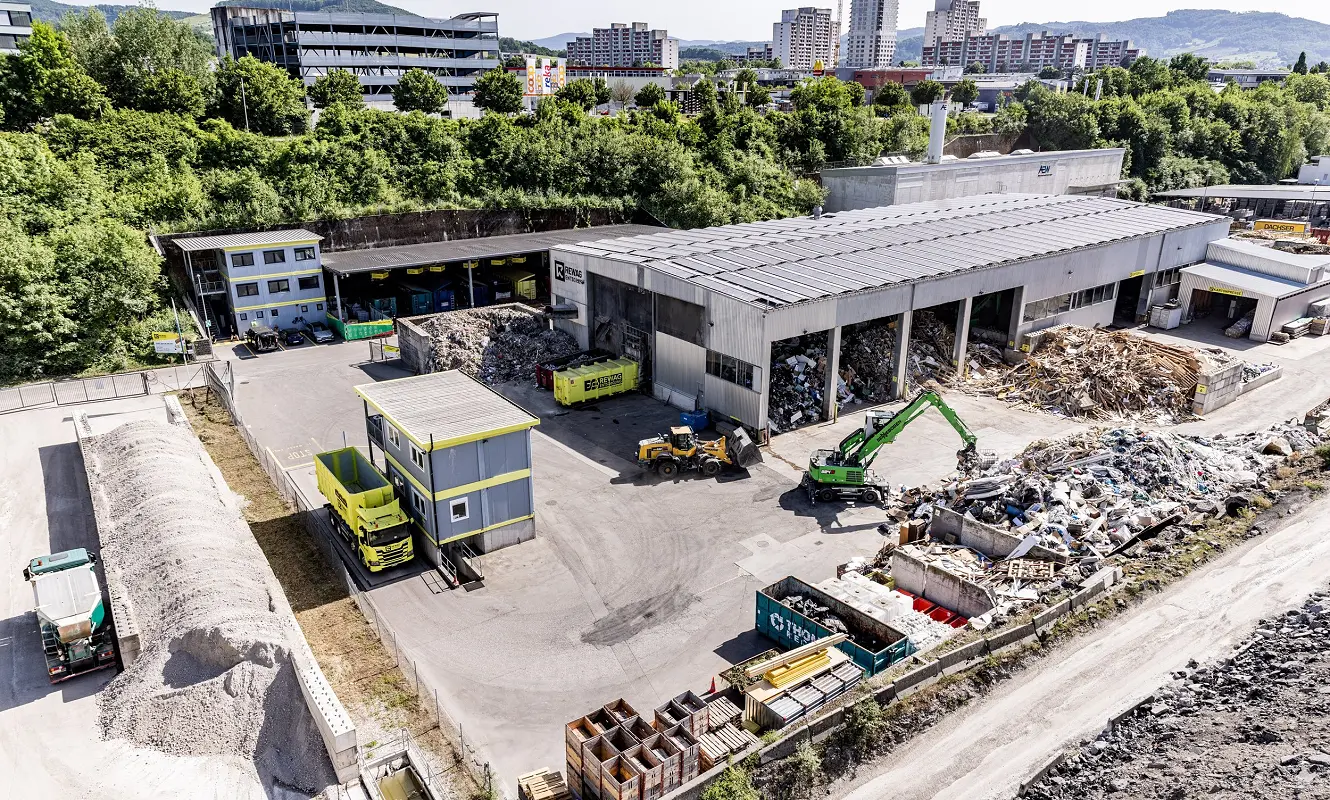 Luftbild des REWAG-Areals mit Sortierhalle, Mulden und Materialhaufen, dahinter die Stadt