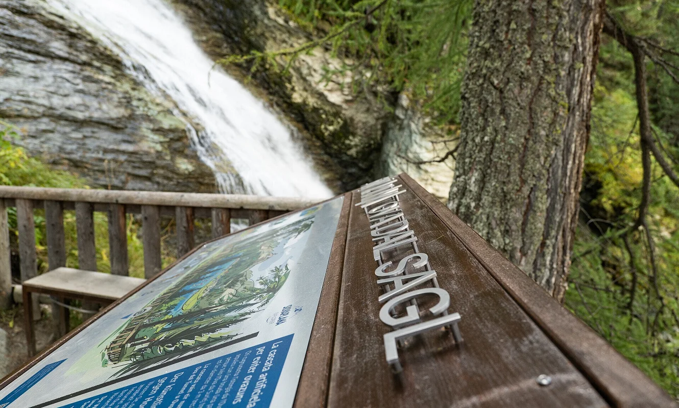 Tafel aus Cortenstahl mit Ortsnamen und illustrierter Karte an einem Holzgeländer beim Wasserfall