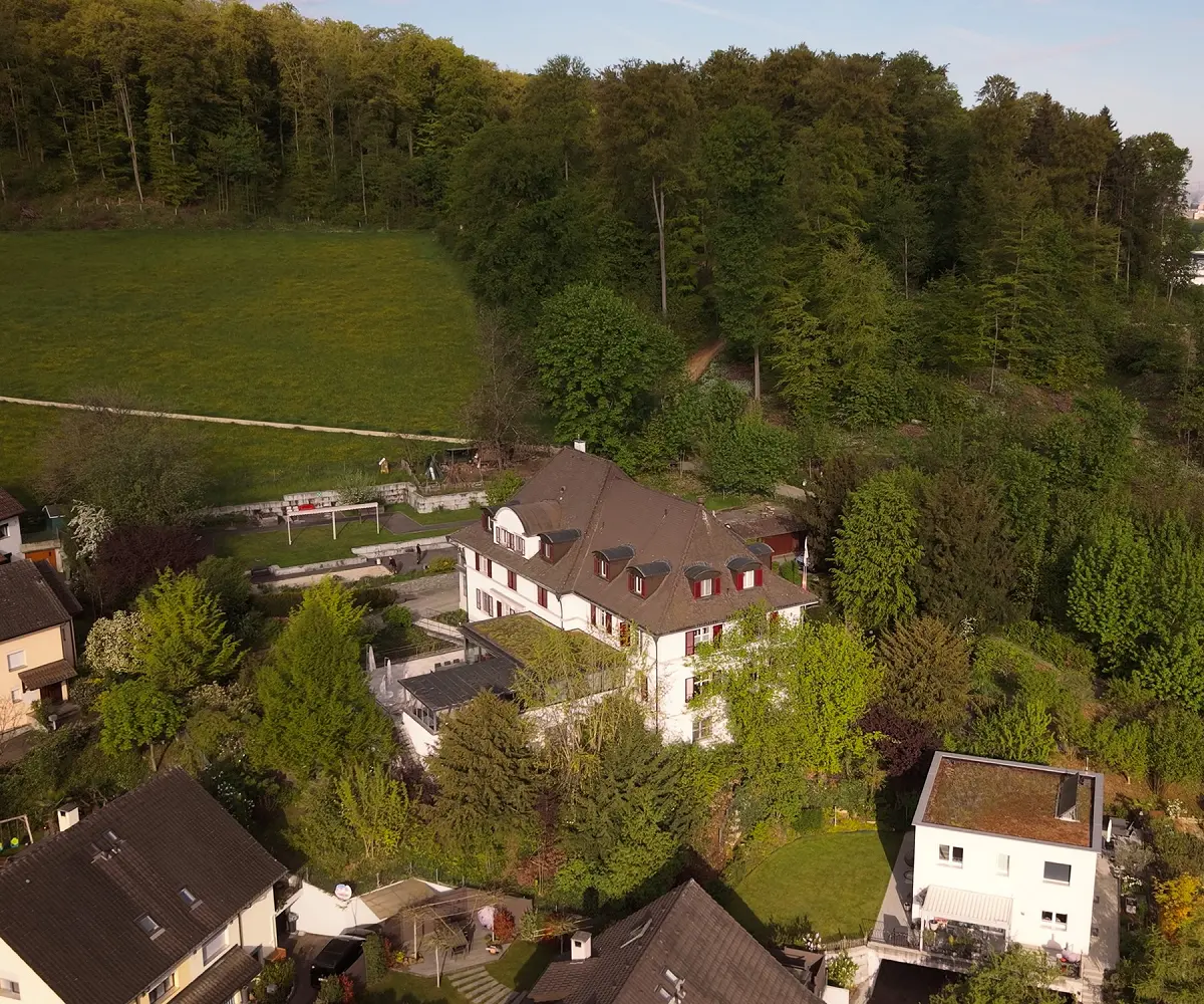 Drohnenaufnahme eines Adulta-Standorts: Villa mit Terrasse am bewaldeten Hang, im Dunst dahinter die Stadt