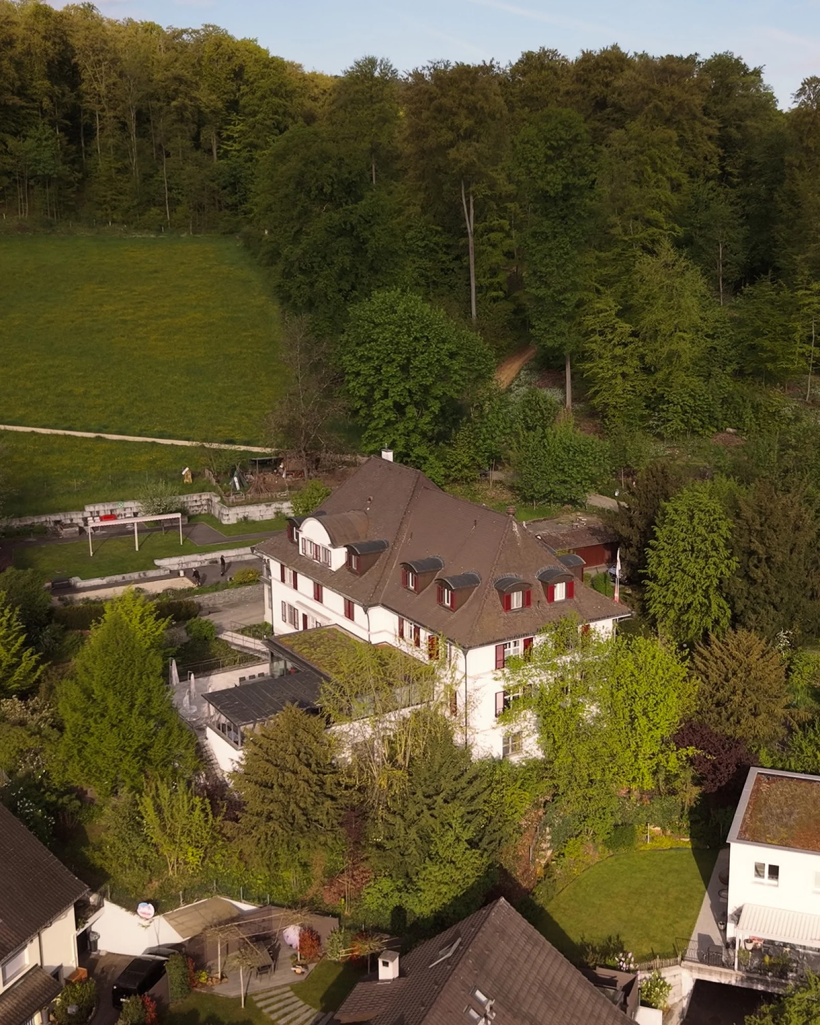 Drohnenaufnahme eines Adulta-Standorts: Villa mit Terrasse am bewaldeten Hang, im Dunst dahinter die Stadt
