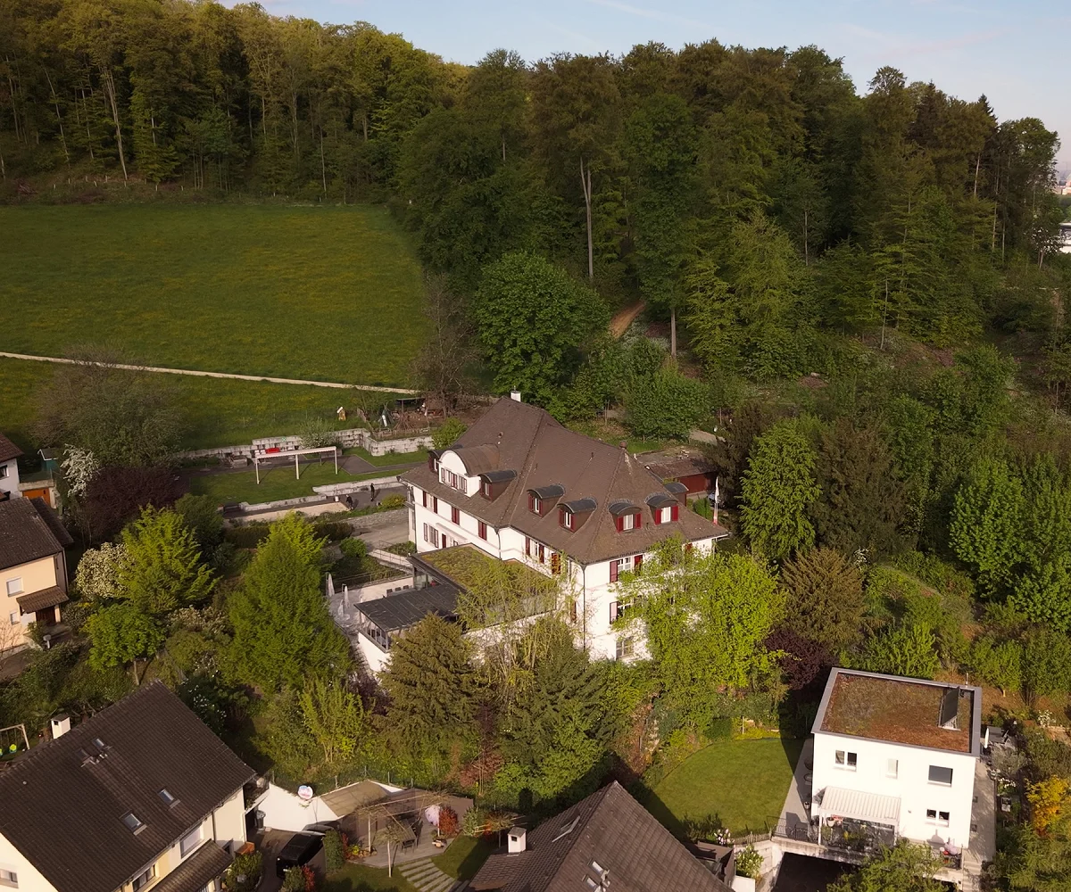 Drohnenaufnahme eines Adulta-Standorts: Villa mit Terrasse am bewaldeten Hang, im Dunst dahinter die Stadt