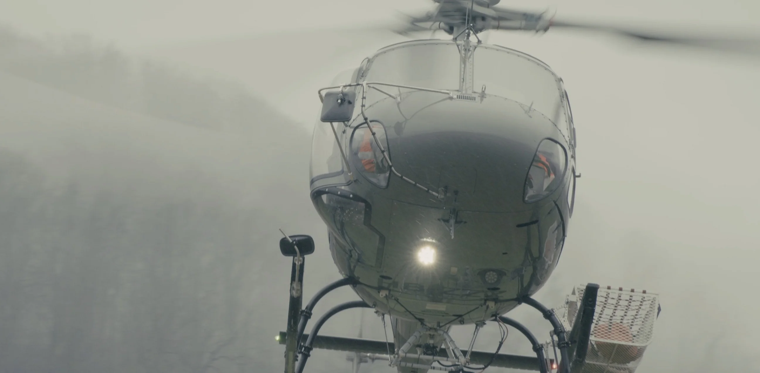 Helikopter im Schwebeflug von vorn, mit Lastseil und Transportkorb, dahinter neblige Bewaldung