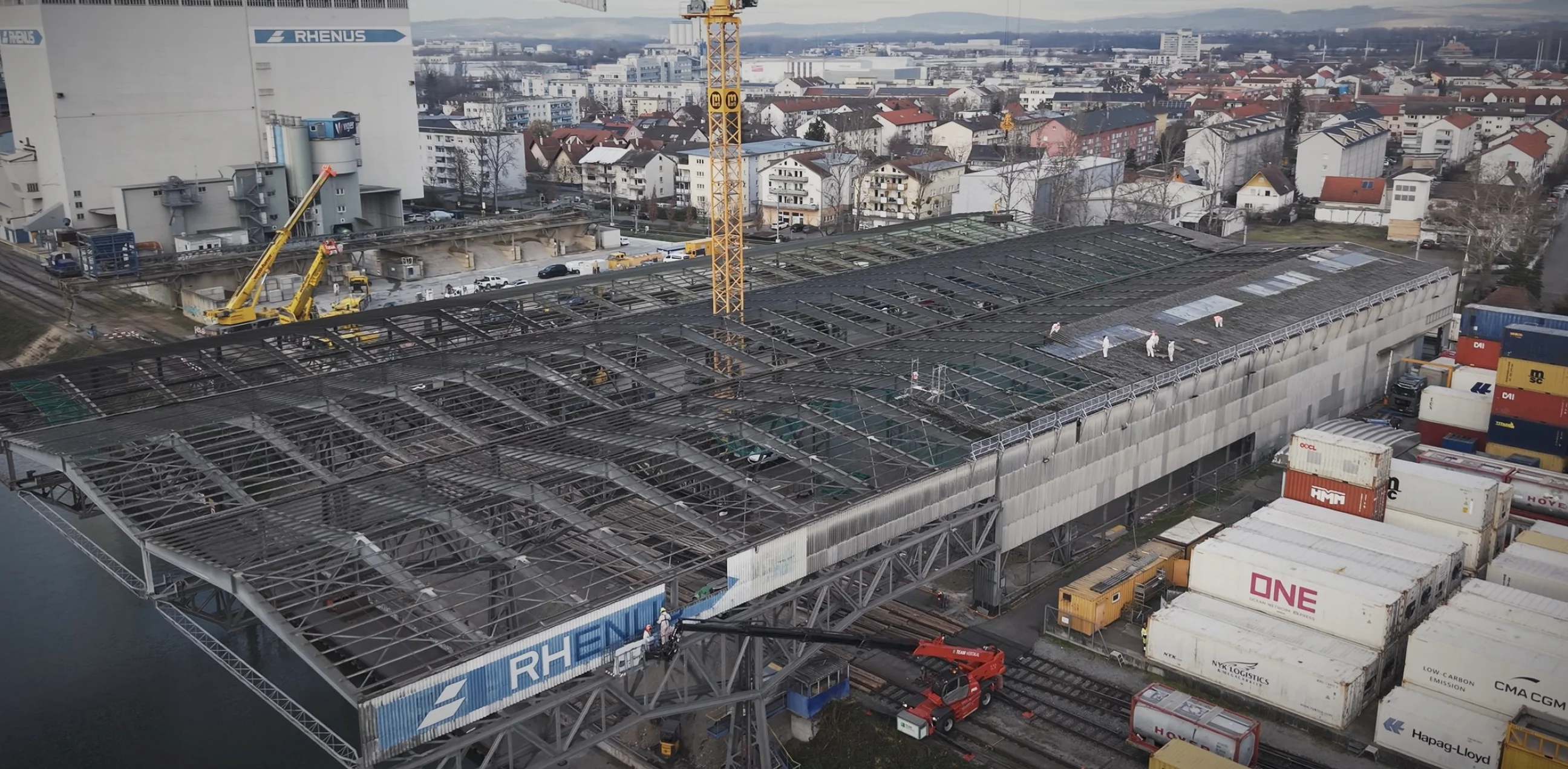 Drohnenaufnahme des Rückbaus: Hallendach bis auf das Stahlskelett abgetragen, Arbeiter in Schutzanzügen, dahinter Rhenus-Silo und Hafen