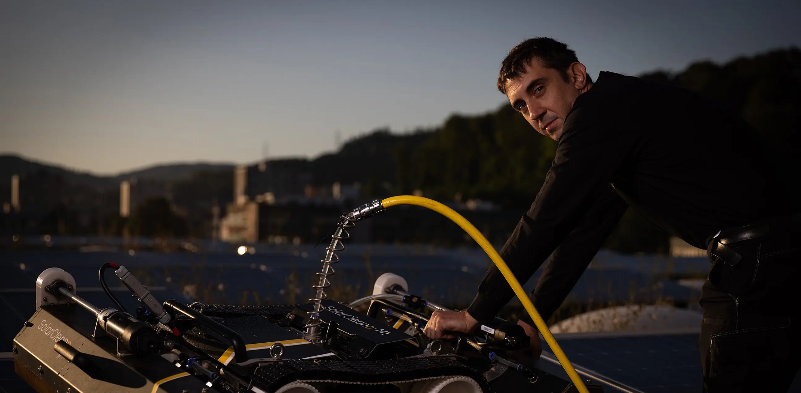 Reinigungsroboter auf einer Photovoltaikanlage, daneben eine Person mit Wasserschlauch in der Dämmerung