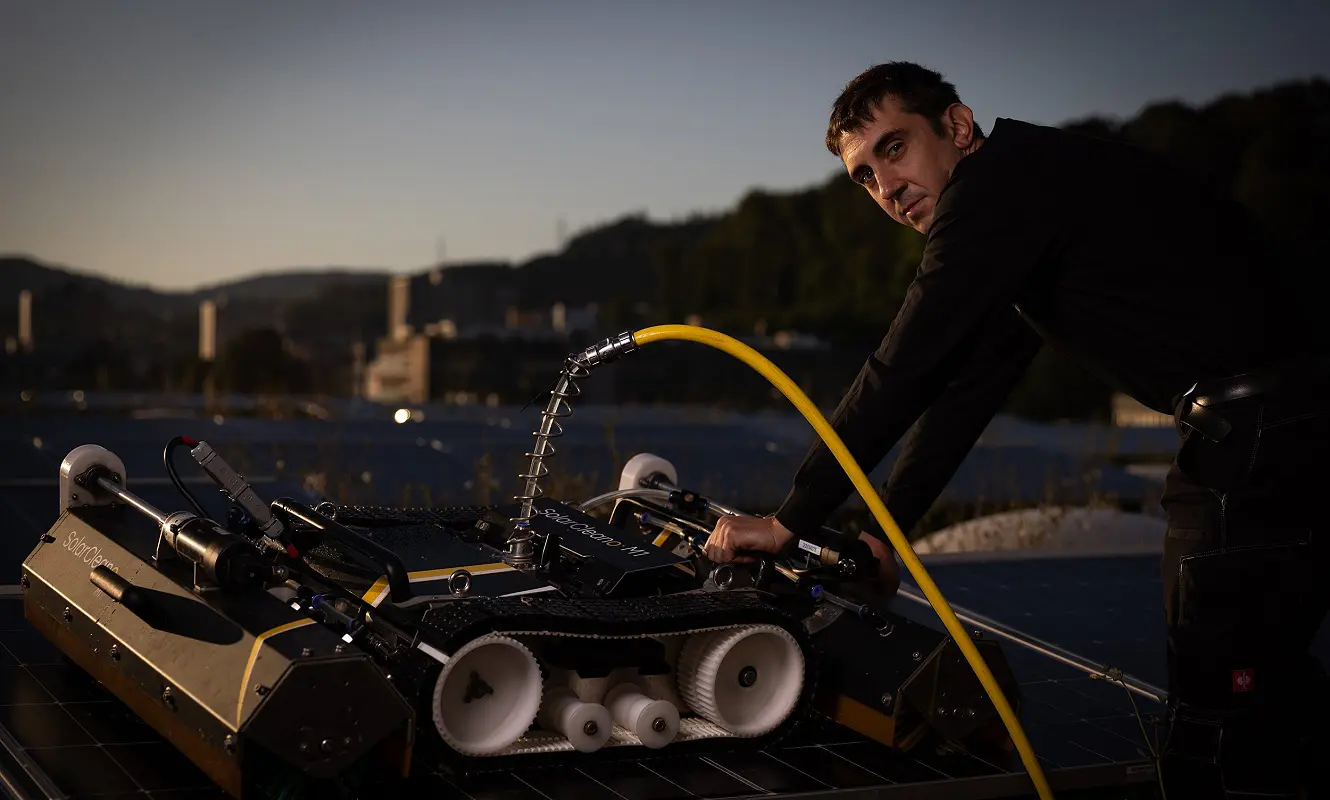 Reinigungsroboter auf einer Photovoltaikanlage, daneben eine Person mit Wasserschlauch in der Dämmerung